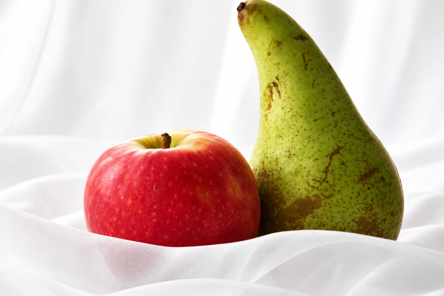 A red apple and a green Conference pear side by side on white fabric.